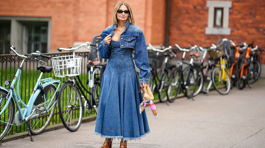 A guest wears black sunglasses, a gold chain necklace, a blue denim micro cropped jacket, a blue denim embossed striped pattern shoulder-off / long ripped dress, a brown shiny leather braided leather Jodie handbag from Bottega Veneta, camel shiny leather varnished platform high heels shoes from Chanel , outside Mark Kenly Domino Tan, during the Copenhagen Fashion Week Spring/Summer 2024 on August 10, 2023 in Copenhagen, Denmark.